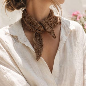May include: A brown knitted neckerchief with a textured ribbed pattern tied in a simple knot around the neck, worn over a long-sleeved white button-down shirt made of a lightweight, semi-sheer fabric with a visible weave and a classic collar.