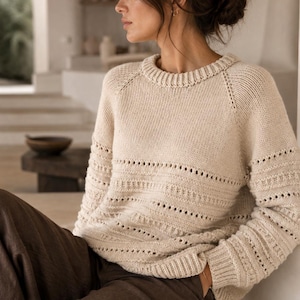 May include: A cream-colored long-sleeve knit sweater featuring a ribbed crew neckline and cuffs, with horizontal bands of openwork eyelet detailing across the chest and sleeves, paired with dark brown trousers.