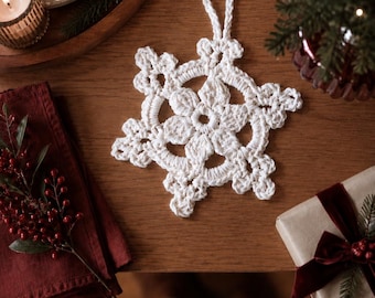 Puede incluir: Un adorno de copo de nieve tejido a ganchillo en color blanco, con un diseño de estrella de seis puntas, patrones calados intrincados y un lazo a juego en la parte superior. Está hecho de hilo blanco grueso con una textura anudada.