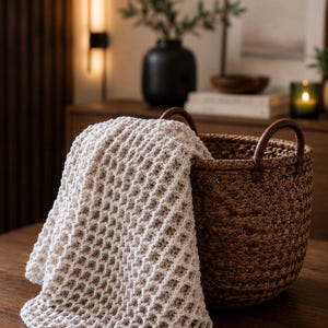 May include: A cream-coloured, waffle-knit throw blanket draped over a round, dark brown woven basket. The basket features two sturdy, curved brown leather handles attached to the rim, providing a contrast to the textured, light-coloured fabric.
