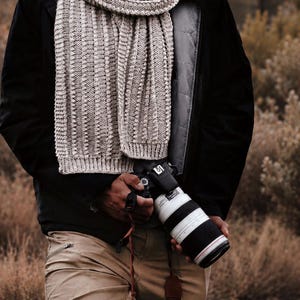 May include: A thick, light gray knitted scarf with a vertical ribbed texture worn over a black jacket and tan trousers. A black camera with a white telephoto lens and a brown leather strap is held in front of the torso, featuring visible lens markings.