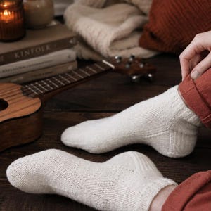 May include: A pair of off-white, ankle-length knit socks featuring a ribbed cuff and a textured, woven pattern. The socks are crafted from a light-colored yarn with subtle flecks, showcasing a classic construction with a reinforced heel and toe.