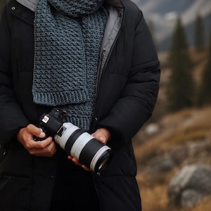 May include: A dark teal chunky knit scarf with a textured pattern worn over a black puffer jacket, paired with a black digital camera featuring a white telephoto lens. The scarf features a ribbed edge and is wrapped around the neck area.
