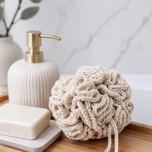 Pode incluir: Uma esponja de banho de crochê na cor creme, de formato redondo, com uma superfície texturizada de laços e um pequeno cordão para pendurar. A esponja é feita de fio grosso e apresenta uma construção densa e franzida para o banho.