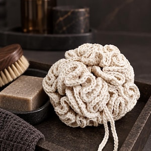 May include: A round, cream-coloured crocheted bath sponge with a textured, ruffled surface and a small loop for hanging. The sponge is crafted from thick yarn and features a dense, layered construction that creates a voluminous, rounded shape.