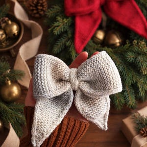 May include: A light gray, hand-knit bow with a textured garter stitch pattern. The bow features two symmetrical loops and two pointed tails, constructed from a soft yarn material. The design is simple, featuring a central knot detail.