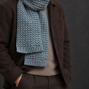 May include: A dusty blue knitted scarf featuring a textured, repeating geometric pattern and ribbed edges. The scarf is draped around the neck, layered over a brown jacket, a neutral-toned sweater, and dark brown trousers, showcasing its length.