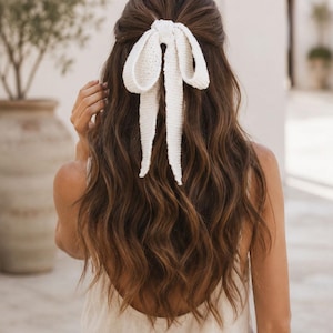 May include: A cream-colored knitted hair bow with long, trailing ribbons secured in a half-up hairstyle. The accessory features a textured, woven fabric construction with a soft, off-white hue, providing a simple and functional way to tie back hair.