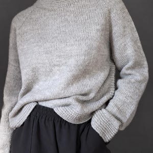 May include: A light gray long-sleeve knit sweater featuring a ribbed crew neckline, raglan sleeves, and ribbed cuffs. The sweater has a textured, heathered appearance and is paired with black trousers that have an elastic waistband.