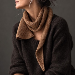 Puede incluir: Una bufanda de punto en color canela con punto bobo y un extremo triangular afilado, envuelta alrededor del cuello. La bufanda se combina con un suéter de punto grueso de color marrón oscuro, manga larga, cuello y puños acanalados.