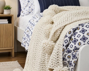May include: A cream-colored chunky knit throw blanket featuring large yarn pom-poms along the edge. The blanket is draped over a bed with white, navy blue, and patterned bedding, including pillows with geometric designs and a solid navy blue lumbar pillow.