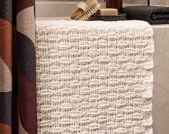 Puede incluir: Una alfombrilla de baño rectangular de color blanco roto con un patrón tejido texturizado de cestería. La alfombrilla está hecha de hilo grueso y muestra un diseño de cuadrícula consistente y repetitivo en toda su superficie, ofreciendo un aspecto estructurado.