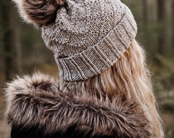 May include: A light beige knit beanie featuring a textured diamond pattern, a ribbed cuff, and a large, fluffy brown and white faux fur pom-pom on top. The hat is worn over long, wavy blonde hair and a black jacket with a faux fur-trimmed hood.