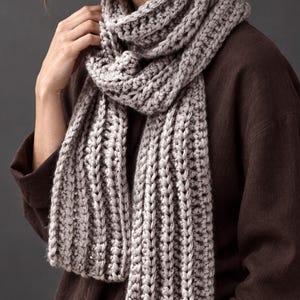 May include: A chunky, light gray knit scarf featuring a prominent vertical ribbed texture. The scarf is wrapped around the neck with long, hanging ends that display the thick, looped yarn construction and consistent stitch pattern throughout.