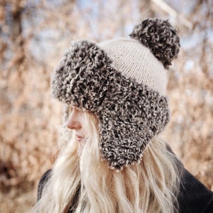 May include: A woman with long blonde hair wearing a white and grey knitted hat with a faux fur pom pom and ear flaps. The hat is worn over a black coat.