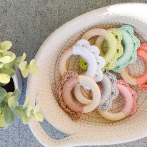 May include: A white woven basket filled with crocheted teething rings in various pastel colors. The rings are made of wood and have a crocheted trim.