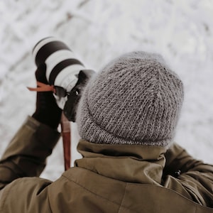 May include: A grey knit beanie worn with an olive green hooded jacket. The individual is holding a professional camera with a white lens, wearing dark brown gloves, and has a thin red strap attached to the camera body for secure handling.