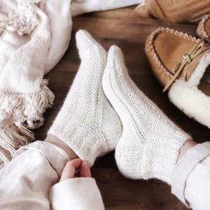May include: A pair of white knitted socks with a textured pattern. The socks are worn by a person sitting on a wooden floor with a white blanket and brown leather pillow in the background.