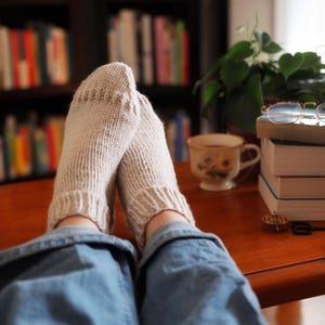 Puede incluir: Primer plano de calcetines de punto beige claro con puños acanalados. Los calcetines descansan sobre una superficie de madera, con una taza de té, libros y una planta en el fondo. La escena evoca una sensación de comodidad y relajación.