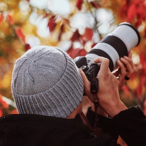 May include: A person wearing a gray knitted beanie and a black jacket is taking a photo with a black and silver camera lens. The background is a blurred image of red and yellow leaves.