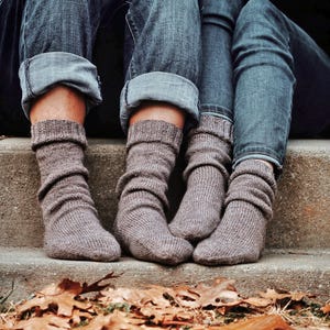 May include: Close-up of three pairs of legs wearing gray knit socks and blue jeans. The jeans are cuffed at the ankles. Brown leaves are scattered on the ground in front of the legs.