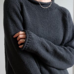 Könnte beinhalten: Ein anthrazitfarbener Langarmpullover mit geripptem Rundhalsausschnitt und passenden gerippten Bündchen. Das Strickkleidungsstück hat eine strukturierte Oberfläche mit sichtbaren Maschen, langen Ärmeln und eine lockere Passform.