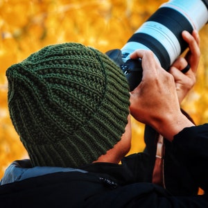 Könnte beinhalten: Eine Person mit einer grünen Strickmütze und einer schwarzen Jacke hält ein Kameraobjektiv und blickt auf einen verschwommenen Hintergrund aus gelben Blättern.