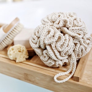 Può includere: Un primo piano di un pouf da bagno in crochet beige con un'ansa a forma di anello. Il pouf è appoggiato su un vassoio di legno con un sapone e una spugna vegetale.