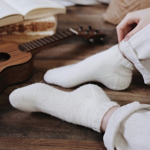 May include: A pair of white knitted socks with a wood floor and a ukulele in the background.