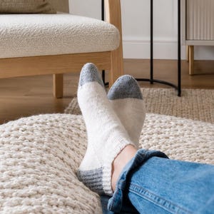 May include: A pair of ankle socks featuring a white main body with contrasting grey toe and heel sections. The socks are worn with blue denim jeans, showcasing a simple knit texture and a snug fit around the foot and ankle area.