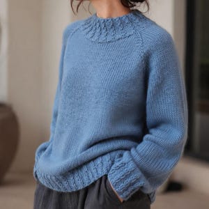 Puede incluir: Un suéter de punto de manga larga en color azul claro con un patrón acanalado texturizado alrededor del cuello redondo y los puños. La prenda tiene un corte relajado con mangas raglán y un acabado de color sólido, combinado con pantalones gris oscuro.
