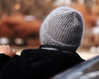May include: A grey knit beanie with a fuzzy, textured surface and a ribbed pattern. The beanie is worn on the head and is paired with a black hooded jacket that features a visible hood and a dark collar, showing the back view of the garment.