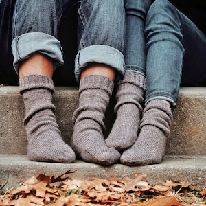 May include: Two pairs of brown knitted socks with cuffed tops, worn by two people sitting on a concrete step with fallen leaves in the foreground.