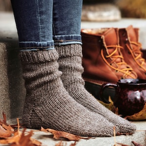 Woman wearing a pair of The Beginner Socks that were knitted using brown worsted-weight yarn. These knitted socks are thick, warm, and cozy, and the knitting pattern is easy to follow and great for beginner knitters to knit a pair of socks.