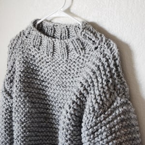 May include: A chunky knit sweater in a light gray color featuring a thick ribbed crew neckline and long sleeves. The garment is crafted with a prominent, textured stitch pattern and is displayed on a white plastic clothes hanger.