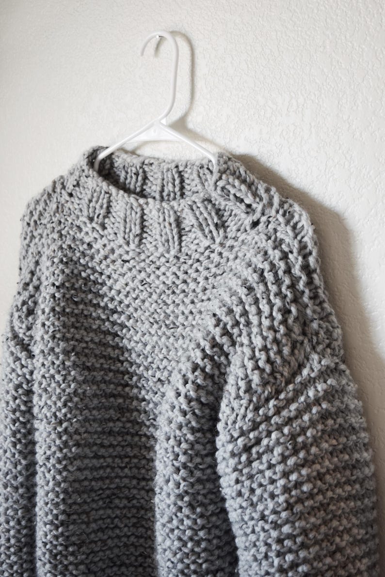 May include: A chunky knit sweater in a light gray color featuring a thick ribbed crew neckline and long sleeves. The garment is crafted with a prominent, textured stitch pattern and is displayed on a white plastic clothes hanger.