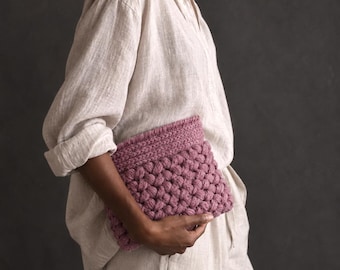 Può includere: Una pochette rettangolare all'uncinetto color malva, caratterizzata da un motivo a punto nocciolina testurizzato sul corpo principale e un bordo a maglia piatta lungo il margine superiore. L'accessorio è realizzato in filato spesso e lavorato a maglia.
