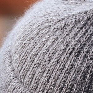 May include: A close-up view of a gray knit beanie featuring a fuzzy, textured surface with visible fibers. The hat is constructed with a standard stockinette stitch pattern and includes a folded brim at the bottom edge, showcasing a consistent gray tone.