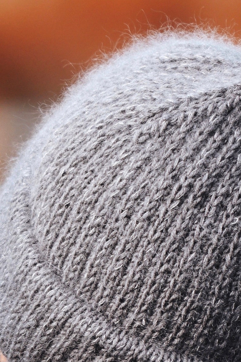 May include: A close-up view of a gray knit beanie featuring a fuzzy, textured surface with visible fibers. The hat is constructed with a standard stockinette stitch pattern and includes a folded brim at the bottom edge, showcasing a consistent gray tone.