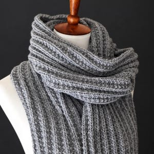 May include: A long, grey knitted scarf featuring a prominent vertical ribbed texture. The scarf is wrapped around a white mannequin neck, displaying a thick, chunky yarn construction with consistent stitch patterns throughout the entire length.