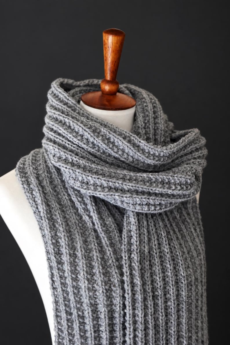 May include: A long, grey knitted scarf featuring a prominent vertical ribbed texture. The scarf is wrapped around a white mannequin neck, displaying a thick, chunky yarn construction with consistent stitch patterns throughout the entire length.