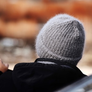May include: A grey knitted beanie with a fuzzy, textured surface and a folded cuff, worn with a black hooded jacket. The beanie features a classic stockinette stitch pattern and a rounded crown, providing a simple and functional head covering.