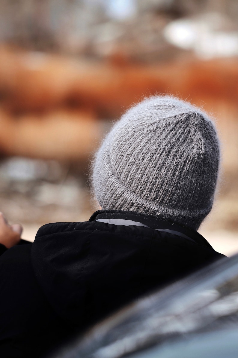 May include: A grey knitted beanie with a fuzzy, textured surface and a folded cuff, worn with a black hooded jacket. The beanie features a classic stockinette stitch pattern and a rounded crown, providing a simple and functional head covering.