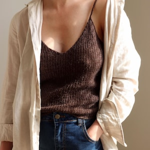 Könnte beinhalten: Ein braunes Strick-Camisole mit dünnen Trägern wird unter einem beigefarbenen Button-Down-Hemd getragen. Das Model trägt eine blaue Jeans. Die Kleidung ist lässig und alltagstauglich.