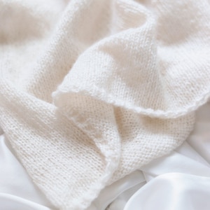 Closeup photo of The Classic Camisole, laid flat and knitted in cream yarn. The easy knit stitches of the tank top are shown, and the fabric of the cami looks soft and lightweight. The bottom of the tank top features ribbing.