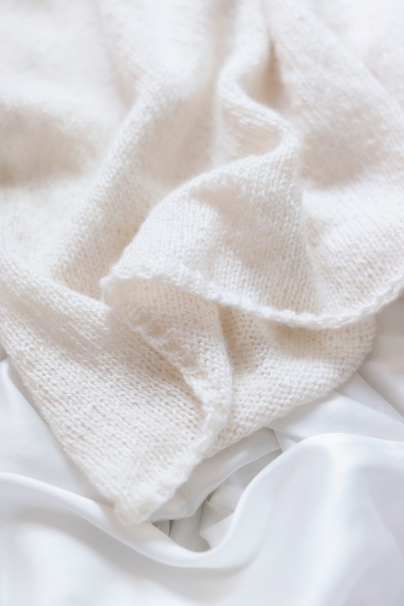 Closeup photo of The Classic Camisole, laid flat and knitted in cream yarn. The easy knit stitches of the tank top are shown, and the fabric of the cami looks soft and lightweight. The bottom of the tank top features ribbing.
