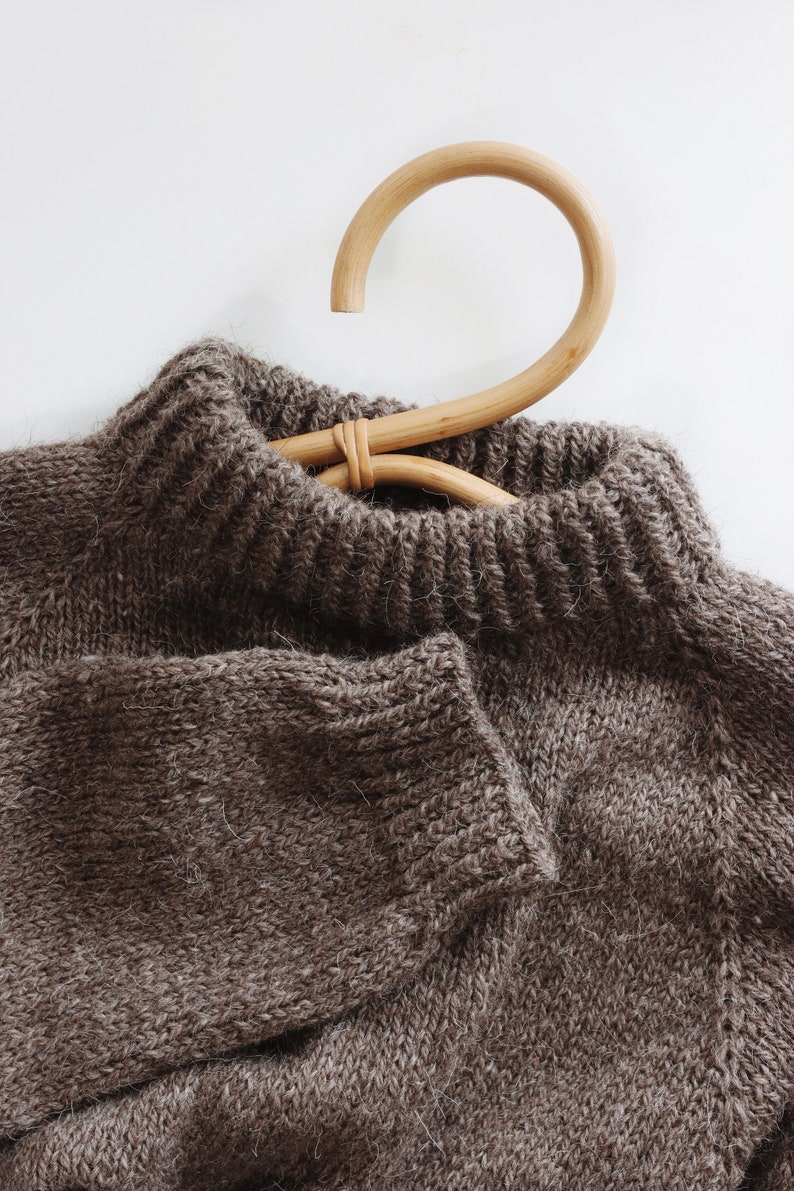 A cozy knitted sweater, The Weekend Sweater, is laid flat knitted in brown yarn. The knit jumper shows off the double-knit neck and ribbing details on the sleeve. This knitting pattern to knit this easy raglan is easy and makes for a cozy sweater!