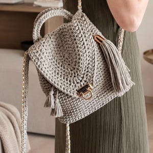 May include: A light gray crocheted backpack featuring a textured stitch pattern, a top handle, and a fold-over flap secured with a gold-toned metal clasp. The bag is accented with matching gray tassels and includes thin straps woven with gold-toned chain links.