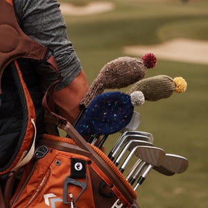 Puede incluir: Tres fundas de punto para palos de golf en marrón, verde oliva y azul marino, cada una con un pequeño pompón. Las fundas están colocadas sobre palos de golf guardados en una bolsa naranja y marrón que contiene varios tees de golf blancos.