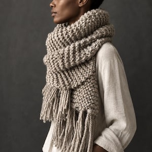 May include: A chunky, beige, hand-knit scarf featuring a thick, textured ribbed pattern and long, twisted fringe tassels at the ends. The scarf is wrapped multiple times around the neck, showcasing a heavy, warm knit construction in a neutral tone.
